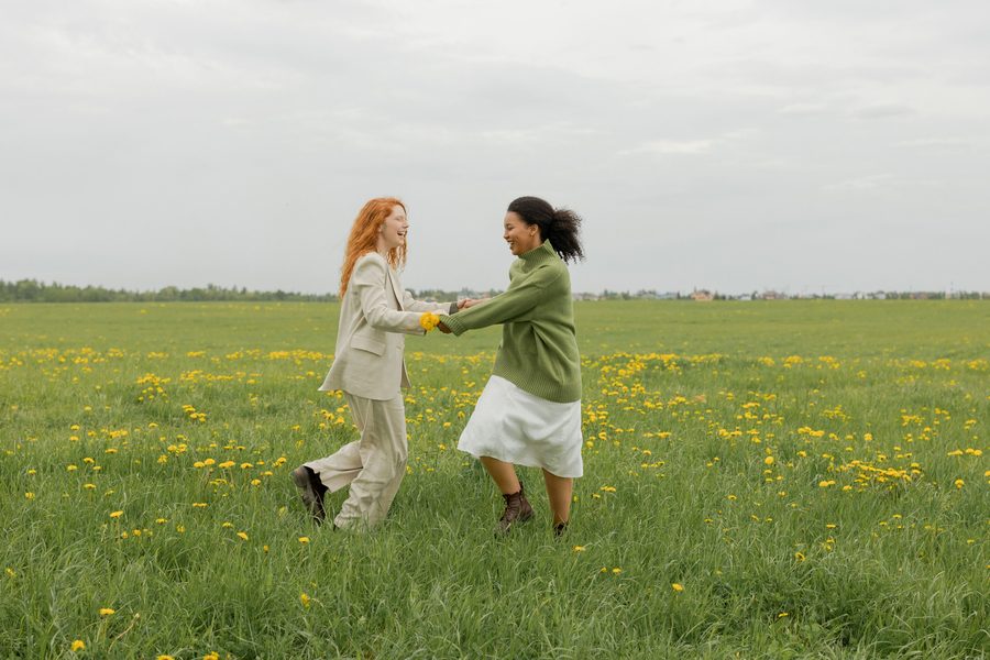 Two women holding hands and laughing in a meadow of dandelions
